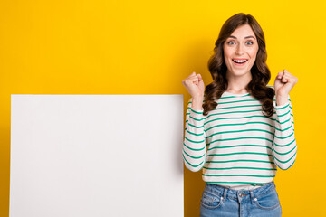 Photo of impressed ecstatic girl with wavy hairdo dressed striped shirt clenching fist near white board isolated on yellow color background