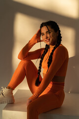 Beautiful young woman with natural make-up wearing orange sports uniform posing near white wall at sunset. Advertising sportswear and yoga wear. Healthy lifestyle, sports