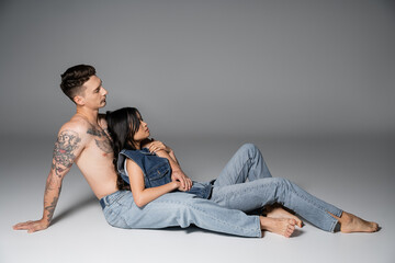 side view of brunette asian woman and shirtless tattooed man in jeans sitting on grey background.