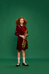 Curly redhaired happy school age girl wearing festive dress playing on saxophone over green...