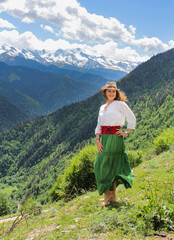 Elegant woman in green skirt at the mountains. Barefoot on the grass. Happy girl with wavy hair with a hat. The background of the majestic mountains of the Caucasus and wooden fence. Svaneti, Georgia.