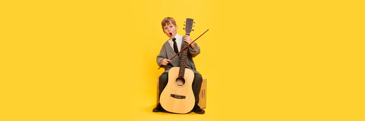 Charming little boy, funny musician wearing huge oversize clothes playing guitar, having fun isolated over yellow background. Pop art, music, new vision, fun concept