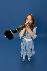Monochrome portrait of charming nice little girl in dress and big shoes playing on clarinet over blue background. Music, fashion, hobby concept - obrazy, fototapety, plakaty