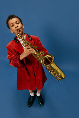 Fototapeta na wymiar Wide angle view of cute little african boy wearing huge man's jacket and shoes like jazz man playing on saxophone over blue background. Fashion, art, music and creative style