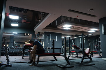 A man with a phone in his hands in the gym. Rest between sets. Dumbbells and kettlebells in the background