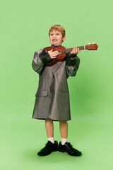 Stylish little school age boy in huge oversize retro style clothes and big male shoes posing with ukulele guitar isolated on green background.