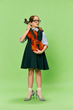 Cute little girl, talanted musician wearing huge mother's heels shoes playing violin, having fun isolated over green background. Education, music, classic art, new vision, fun concept