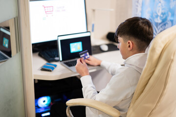 Fototapeta premium Happy teenage boy using laptop and credit card, sitting at desk and ordering goods online from home.