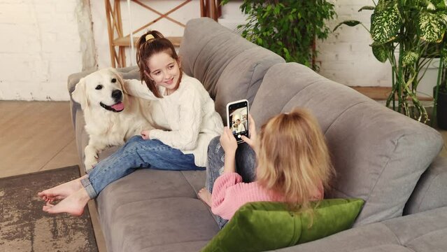 Two little girls, kids playing on sofa with purebred dog, golden retriever and taking photos on phone. Memories. Concept of family, childhood, pets, care, friendship, emotions. leisure