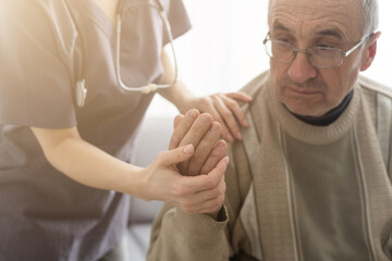 Caring nurse talks to old patient holds his hand sit in living room at homecare visit provide...