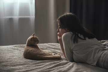 A beautiful young girl in pajamas lies on a bed at home, next to her peach British cat