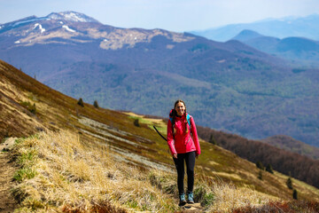 Fototapeta premium girl in pink jacket walks along a mountain ridge during spring; mountain walk with vegetation coming to life, colourful spring in the mountains