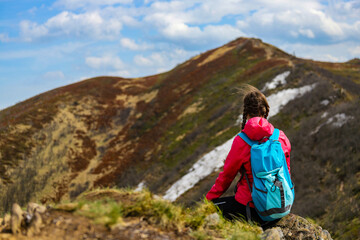 beautiful hiker girl with backpack sitting on the grass with large colorful mountain in front of...