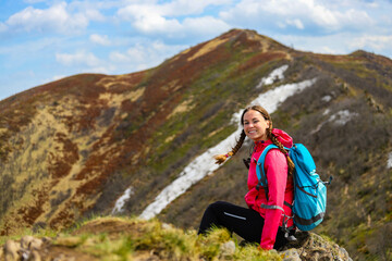 Naklejka na ściany i meble beautiful hiker girl with backpack sitting on the grass with large colorful mountain in front of her; hiking in the mountains during early spring
