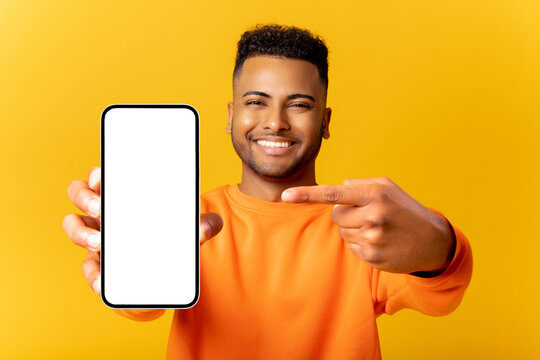 Surprised Indian man holding smartphone in hand and points finger at empty phone screen, astonishment guy presenting best deal, advertising new mobile app, isolated on yellow