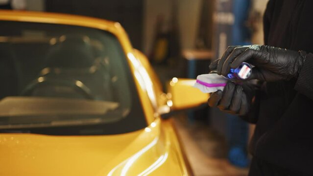Cropped image of hands of male auto service worker in protective gloves, applying special polish cream, nano-ceramic coating on sponge, to polish yellow luxury car on the background. Car detailing