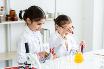 Two little Asian girls in white lab coat help each other for experiment. Little glasses girl using pipette drop solution in yellow chemical flask and her friend using tablet for recording information.