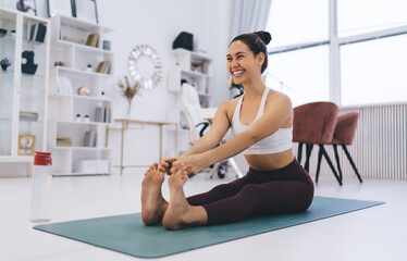 Obraz premium Smiling young woman doing seated forward bend yoga in living room