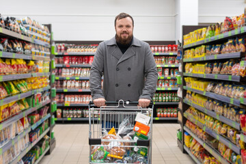 Smiling, contented young man with a beard and a gray coat is standing in a grocery supermarket with a shopping cart filled with food