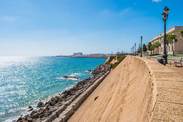 Stone wall and breakwater with blue sea and coastline of Cádiz with sunlight on horizon, SPAIN © Liliana