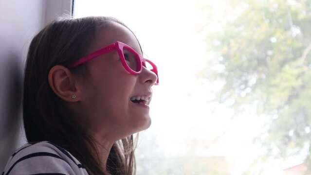 a girl is sitting on the window and laughing,it's raining outside,a child in pink glasses,a joyful preschooler
