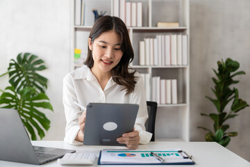 Attractive asian businesswoman in office holding tablet working with laptop and with documents at her desk checking business documents, auditing, searching documents, tax, accounting