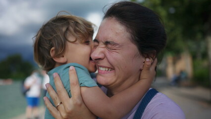 Funny cute moment of child kissing mother in cheek with tongue. Real life authentic family motherhood concept. Little boy bonding with parent outdoors
