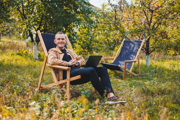 Smiling young freelancer man sitting in wooden chair with laptop while working outdoors in garden. Remote work.