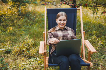 Young woman in a checkered shirt sitting in a chair outdoors in a garden and working on a laptop, remote work, female freelancer
