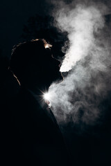 Dark photo of a silhouette of a man blowing smoke against the light. Black background and white...