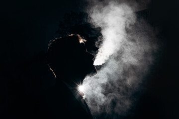Dark silhouette of the face of an adult man blowing smoke against the light. Smoking in the dark. Bad habits of adults people