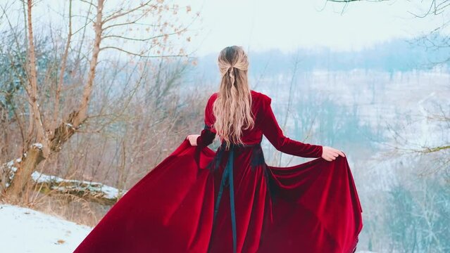 Fantasy portrait happy young woman stands on top of mountain holds skirt dress hands, red fabric flies waves in wind. Princess girl enjoys winter nature mountains snow falls. Lady back rear view. 4k