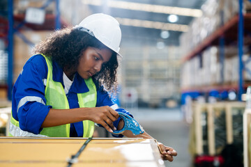 Young beautiful female worker wearing Safety helmet using tape dispenser gun while packing cardboard boxes for the shipment in a distribution warehouse.