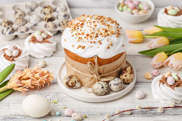 Traditional easter cake or sweet bread, quail eggs, white meringues in shape of nest, pussy willow twigs and spring flowers over white wooden table. Side view. Easter treat, holiday symbol