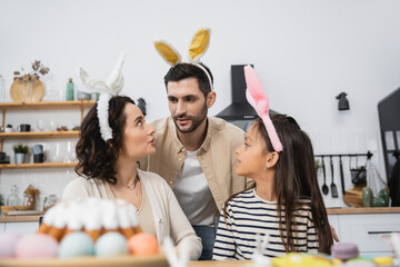 Family in bunny ears headbands sitting near blurred Easter Cake in kitchen.