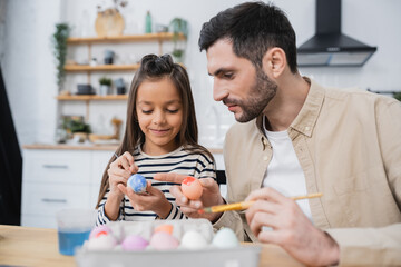 Smiling girl painting Easter eggs with father in kitchen.
