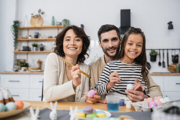 Fototapeta premium Smiling family looking at camera while coloring Easer eggs in kitchen.