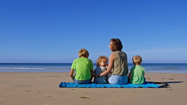 Mom with three kids on mat sit at ocean beach and turn back
