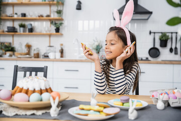 Obraz na płótnie Canvas Smiling child in headband looking at Easter cookie near food in kitchen.