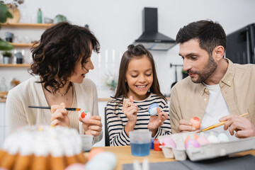 Smiling child coloring Easter egg near parents and blurred cake at home.