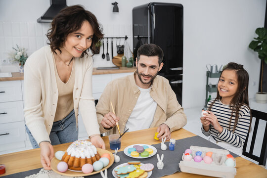 Family coloring Easter eggs near cheerful mother holding plate with cake at home.