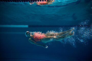 Swimming on back. Professional female swimmer training in swimming pool indoor. Cap and goggles. Underwater view. Concept of sport, endurance, competition, energy, healthy lifestyle
