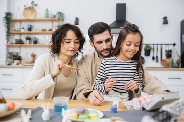 Parents and smiling kid holding paintbrushes near Easter eggs in kitchen.