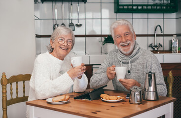 Fototapeta na wymiar Happy senior couple having breakfast together at the table at home looking at camera smiling. Two old people enjoying retirement together