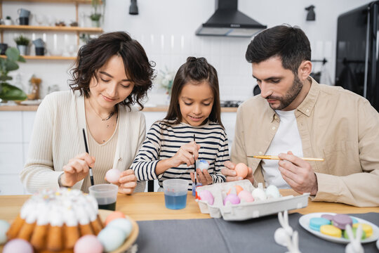 Family painting Easter eggs near blurred cake on table in kitchen.