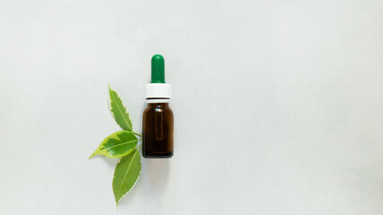 Brown glass bottle with dropper cap. Bottle for cosmetic oil. Minimalist decor with three green fresh leaves. Light gray background. Close up, copy space. body care concept