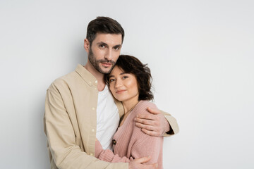 Bearded man hugging wife and looking at camera on white background.