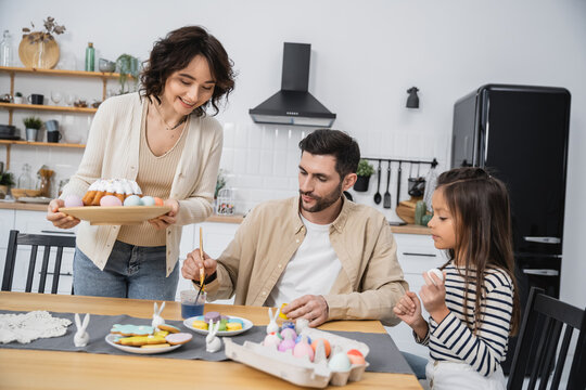 Positive woman holding plate with Easter cake while family coloring eggs at home.