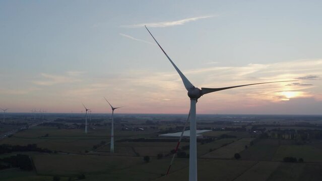 Aerial shot by a drone of windmills with rotating wings among green fields against a sunrise or sunset background. Wind farm with turbine cables for wind energy. Renewable energy source, earth care