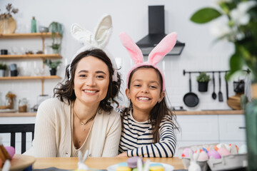 Smiling mom and kid in Easter headbands looking at camera near eggs and macaroons at home.